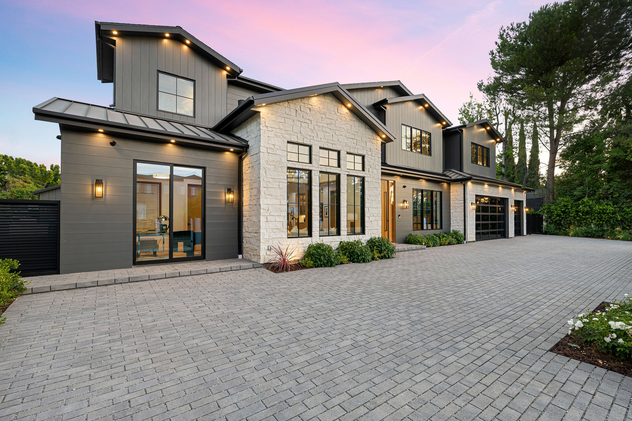 Modern luxury home exterior with large windows, stone facade, and spacious driveway at sunset.